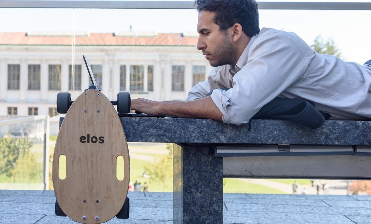 An Elos board leaning against a stone bench while its rider works on a laptop